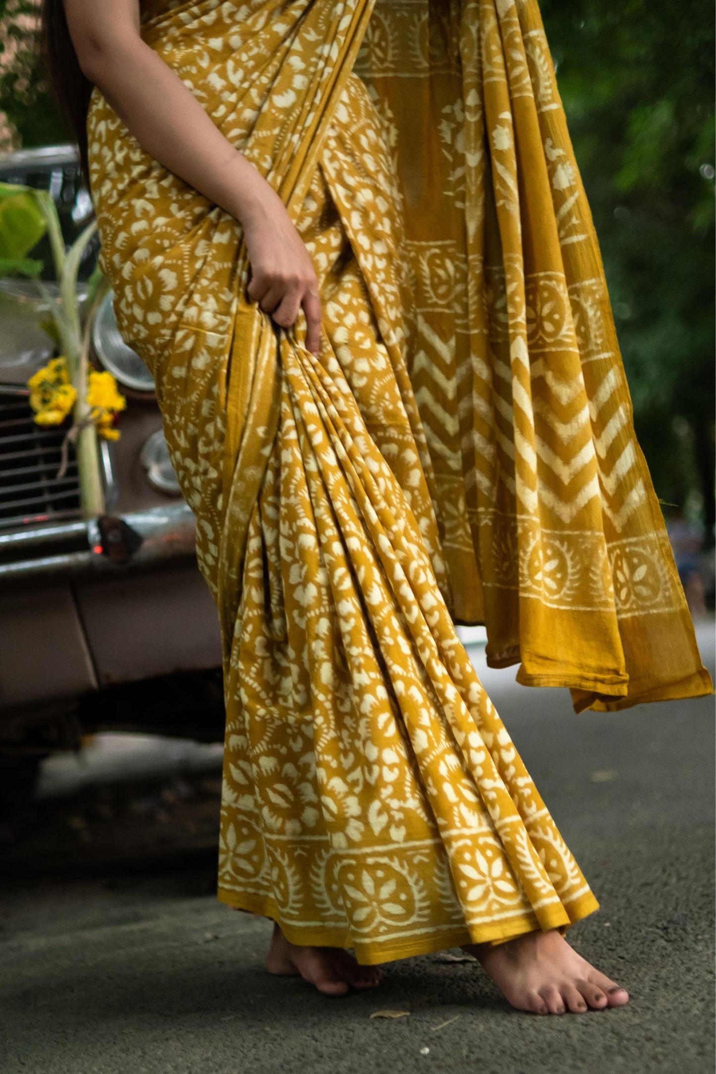 Person wearing a yellow saree with a pattern, standing on a road.