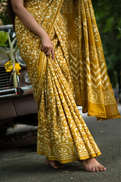 Person wearing a yellow saree with a pattern, standing on a road.